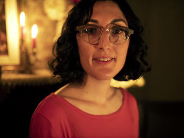 A Middle Eastern woman with light skin and dark hair, wearing glasses and a red shirt, smiling.