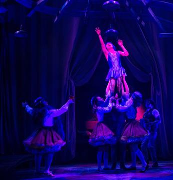 Three actors hold up another performer, who reaches toward the ceiling as they sing. Two other actors look on.