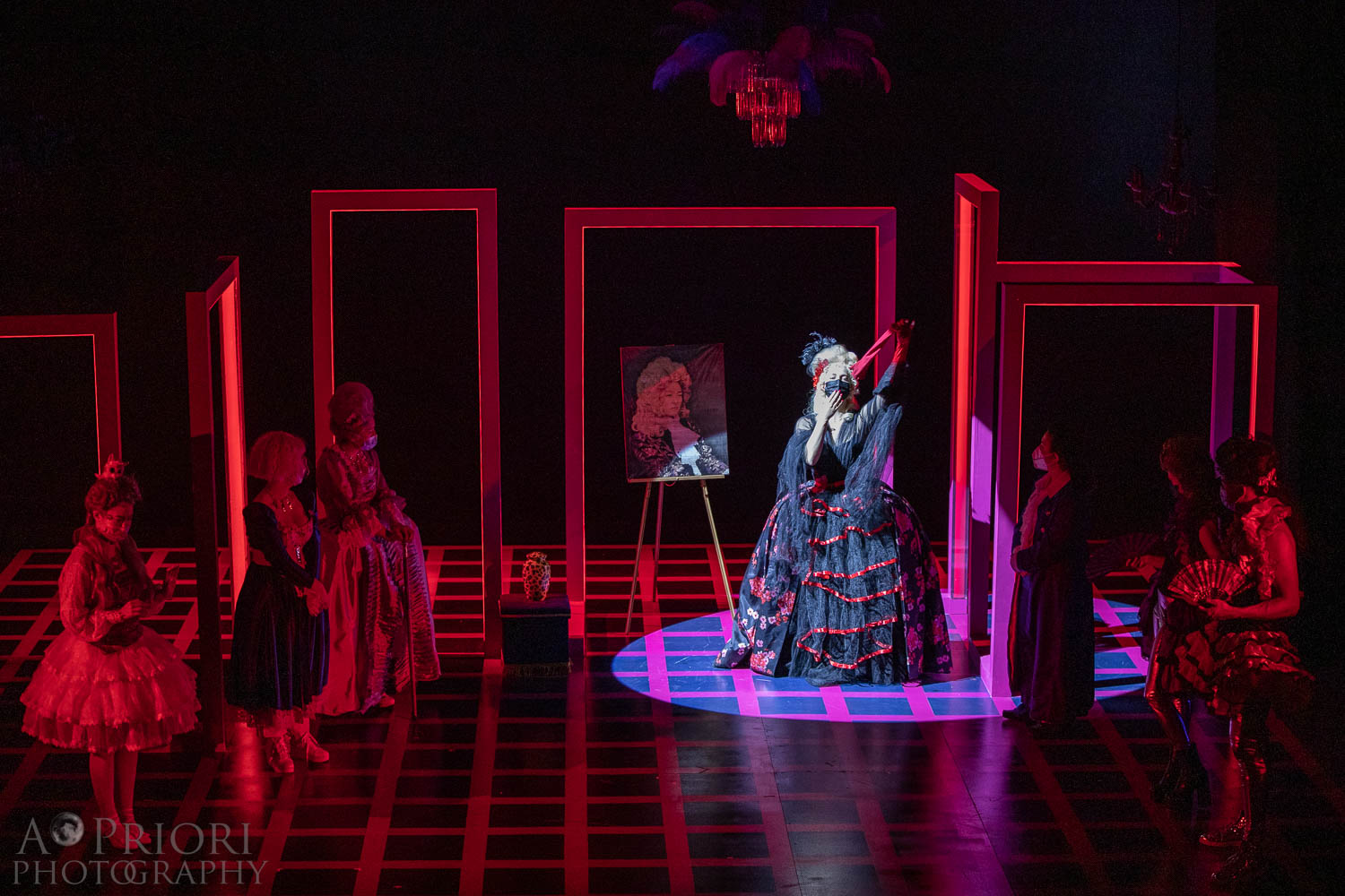 A spotlit performer in a black and red dress reminiscent of Restoration era fashion pulls off a red glove dramatically. The performer stands next to a large portrait of a serious person dressed in Restoration era wear. Other performers look on.