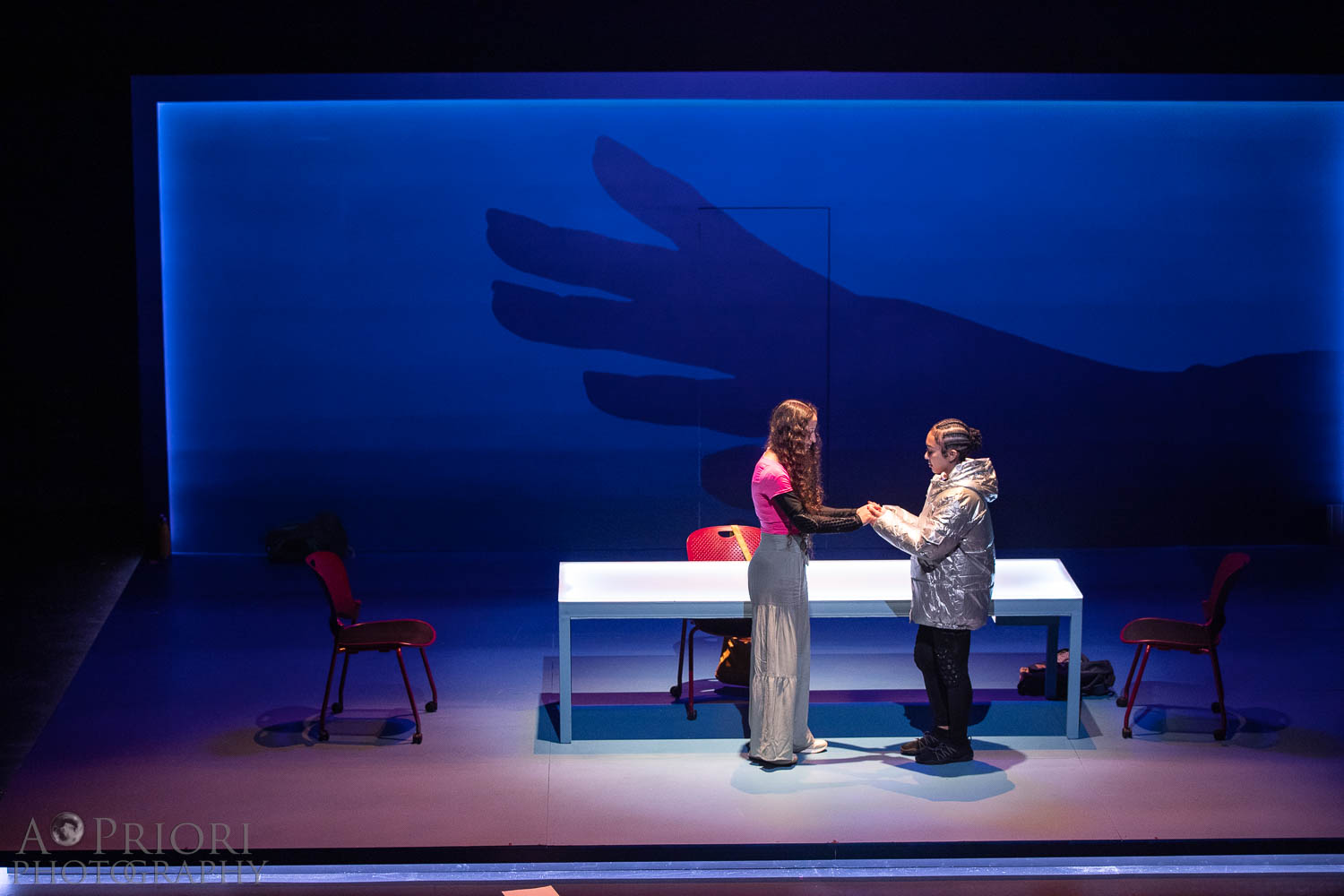 Two performers stand together, clasping hands. Projected on the wall behind them is the shadow of a hand reaching out